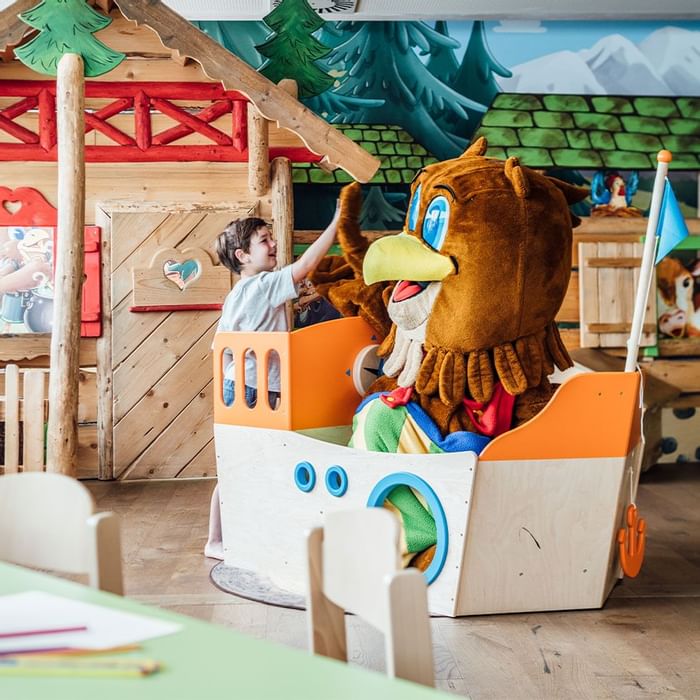 Child inside a wooden play boat with a mascot in the play area at Falkensteiner Hotel Sonnenalpe