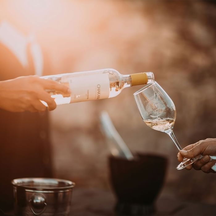 Due mani versano vino bianco da una bottiglia in un calice da vino all'aperto.