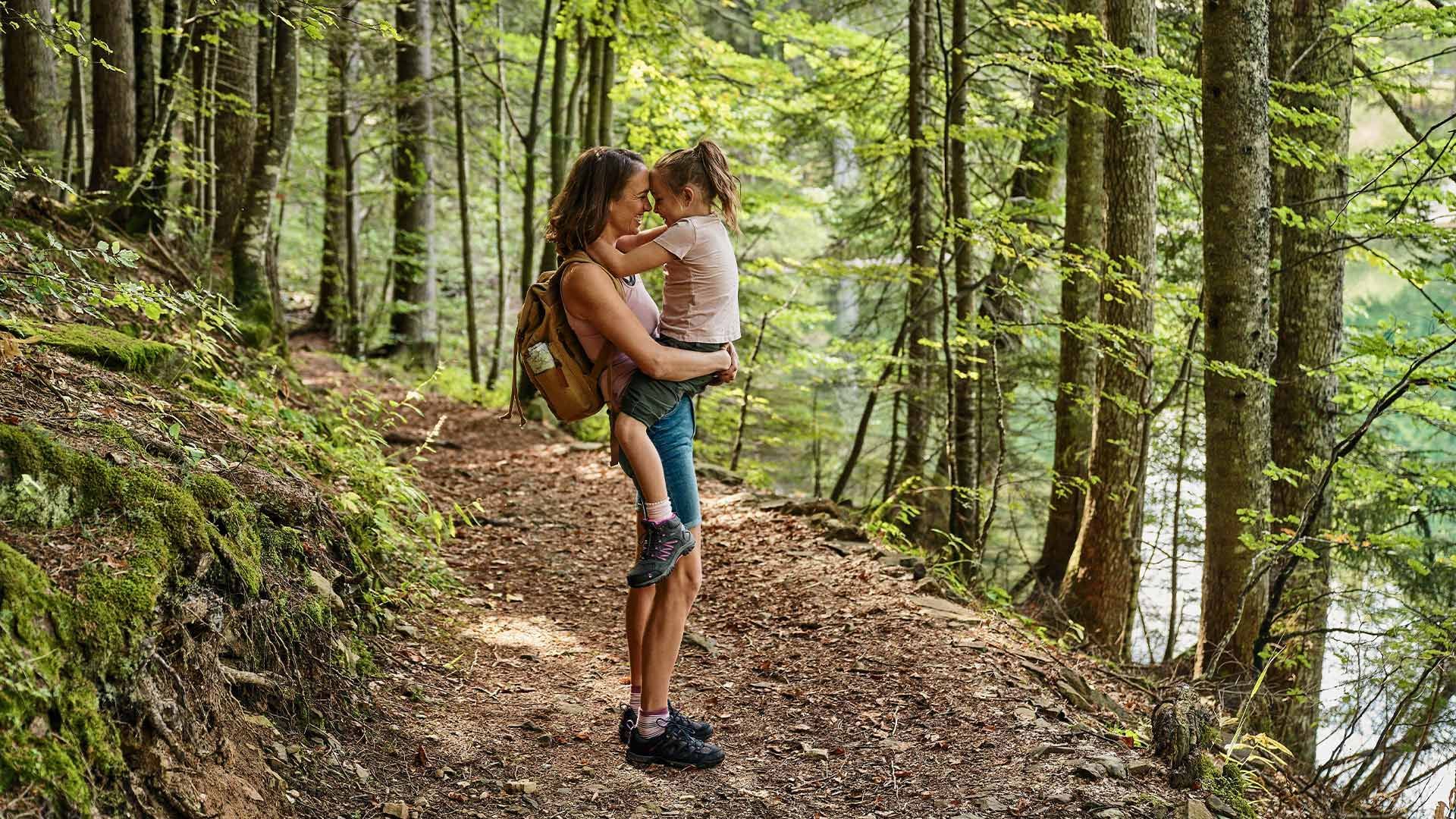 Woman with a child on her back hiking on a dirt trail surrounded by lush forest.