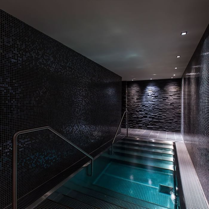 Piscina interna illuminata con rivestimento in mosaico nero e gradini metallici.